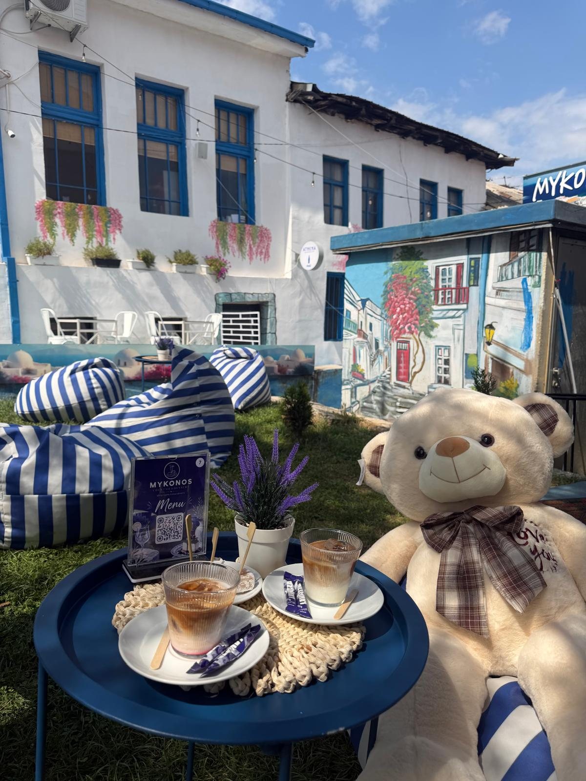 Two iced coffees on a blue tray table beside a giant teddy bear, murals of a Cycladic lane behind